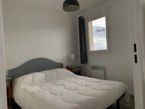 A bedroom with a bed, a nightstand, a lamp, a window with a view of snow-covered mountains, and a ceiling light. - Charming Ski-in/Ski-out Apt with Balcony, Sleeps 6, Access to Spassio Spa, Close to Amenities (Peyragudes)