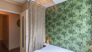 Quarto com cama, luminária pendente, porta e parede com papel de parede tropical.
