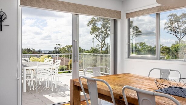 A modern interior with a wooden dining table and white chairs, large windows with a view of trees and a distant tower, and a balcony with outdoor seating.