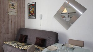 A modern living room with a grey sofa, patterned throw, a poster, a wall-mounted mirror, and a dining area with a glass table and chairs. - Renovated well-equipped studio, south exposure, Chamrousse (Chamrousse)