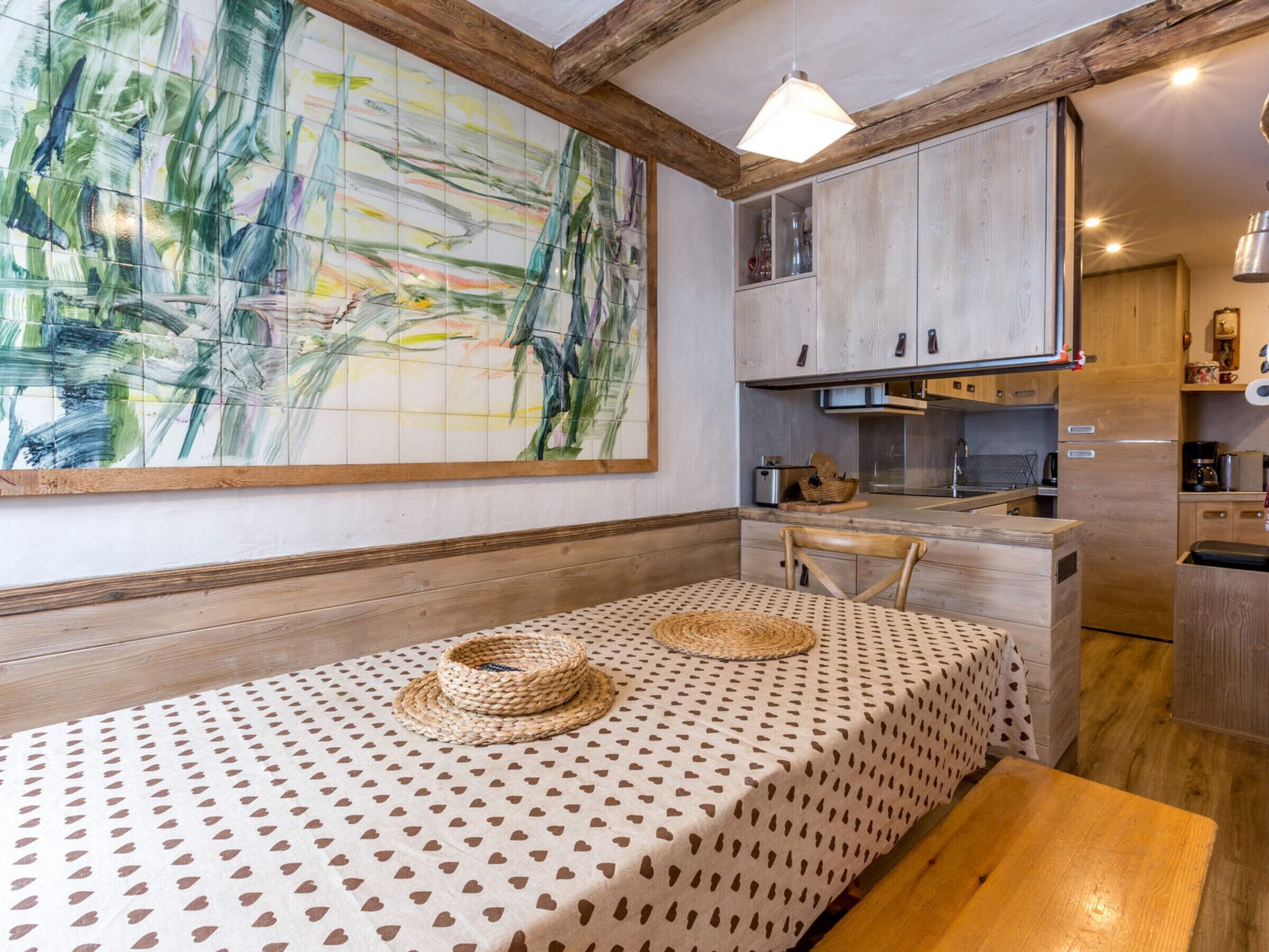 Una cucina compatta con mobili in legno, un tavolo da pranzo con tovaglia fantasia e un grande quadro astratto appeso alla parete.