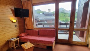 Una habitación con pared de madera, un televisor sobre un sofá, una mesa pequeña con un vaso, y vista a un paisaje montañoso a través de un ventanal grande.