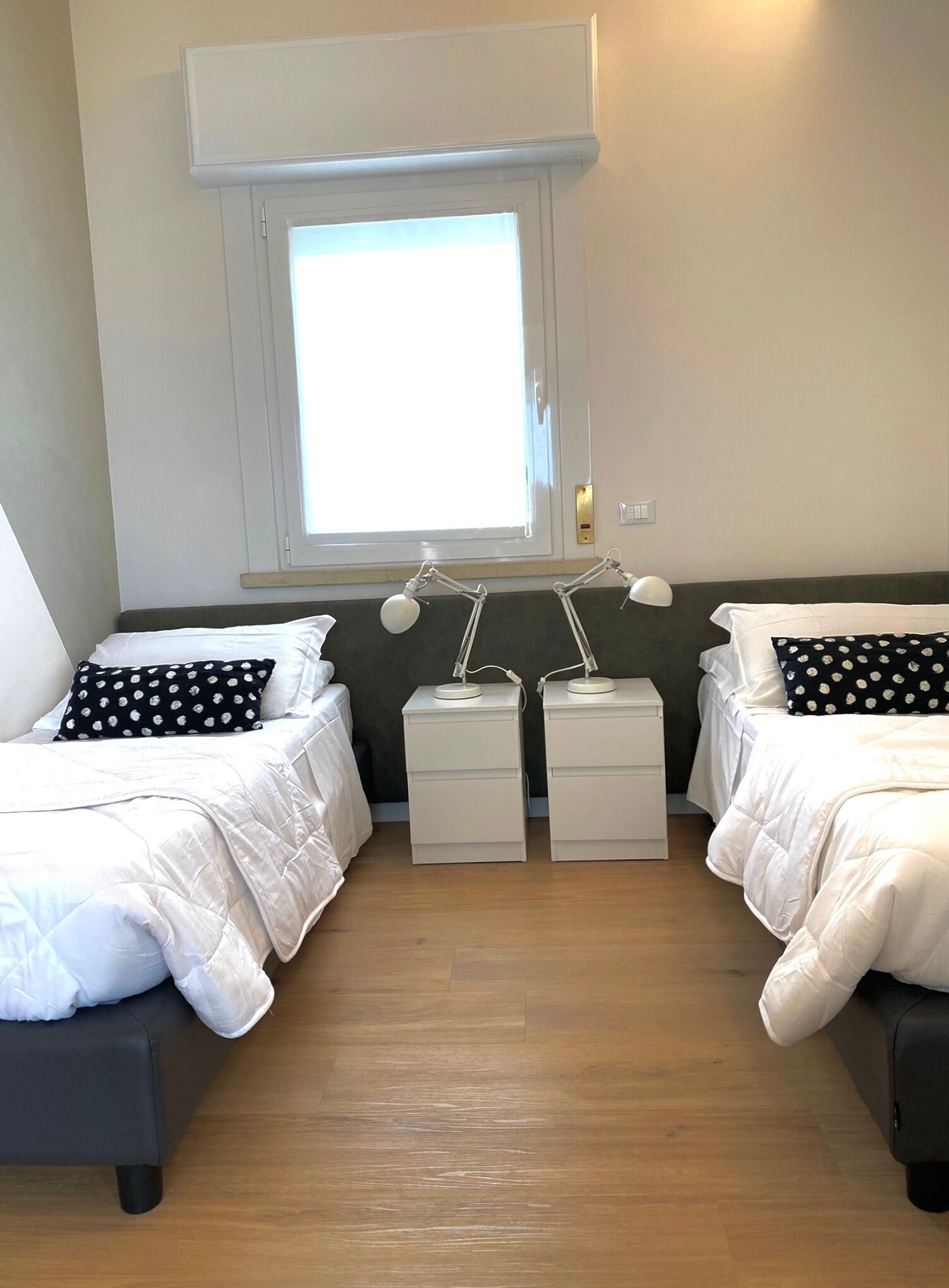 A bedroom with two beds, each with white bedding and black pillows with white polka dots. There are bedside tables with lamps, a window with blinds, and a light switch on the wall.