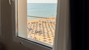 Blick durch ein Fenster auf einen Sandstrand mit vielen Sonnenschirmen und Liegestühlen.
