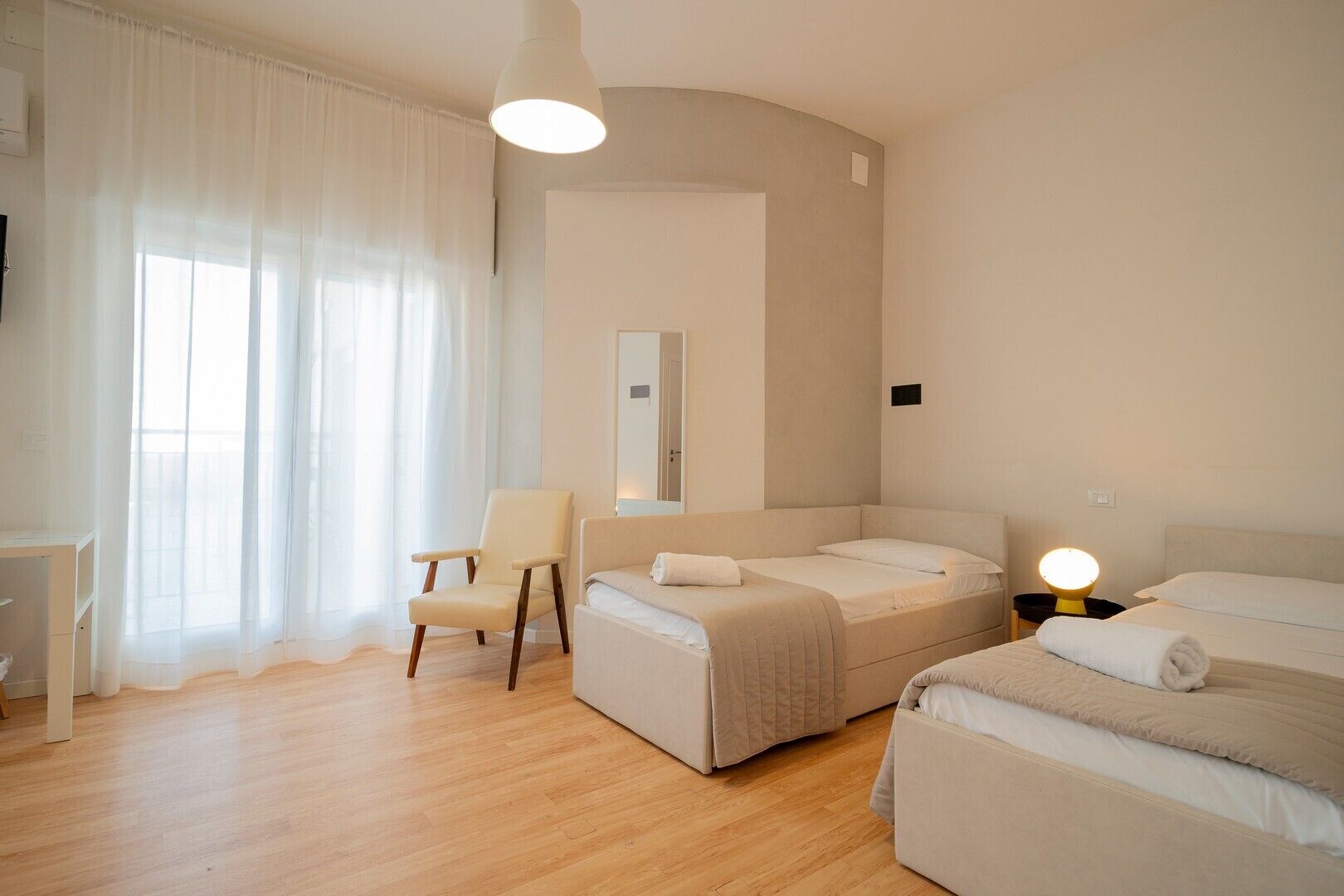 Une chambre d’hôtel moderne avec deux lits, une chaise, un bureau et une grande fenêtre agrémentée de rideaux légers.