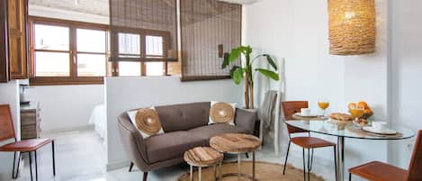 Una sala moderna con sofá, mesa de centro y sillas. Se observa un comedor con una mesa y sillas. También se distingue una planta grande y una lámpara de techo tejida.