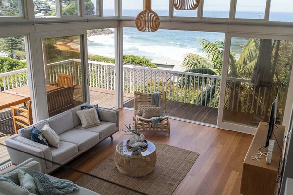 A spacious living area with a large window overlooking the ocean, a wooden deck, and a cozy seating arrangement. - 'The Bungan' - Absolute Beachfront, Direct Beach Access to Bungan Beach, Newport (Newport)