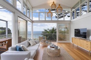 A spacious living room with a large sofa, a coffee table, and a TV on a wooden cabinet. There are large windows offering a view of the ocean and a balcony with a railing. - 'The Bungan' - Absolute Beachfront, Direct Beach Access to Bungan Beach, Newport (Newport)