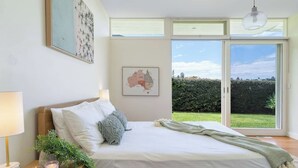 A bedroom with a large bed, a nightstand with a lamp, a map on the wall, and a view of a green garden through the window. - 'The Bungan' - Absolute Beachfront, Direct Beach Access to Bungan Beach, Newport (Newport)