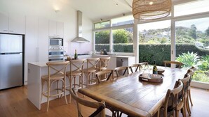 A modern kitchen with white cabinetry, a central island, and a dining area with wooden chairs. - 'The Bungan' - Absolute Beachfront, Direct Beach Access to Bungan Beach, Newport (Newport)
