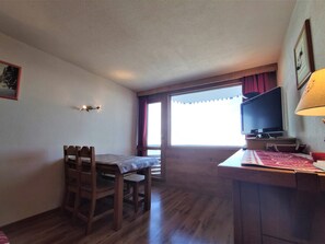 Um quarto com uma mesa de jantar de madeira e cadeiras, uma televisão em um suporte e uma janela com cortinas.
