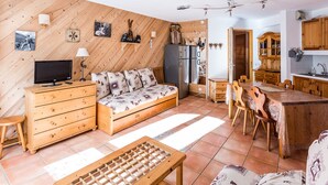 Un salon chaleureux et bien éclairé, avec un canapé, un téléviseur, des meubles en bois et un coin cuisine doté de placards en bois et d’une table à manger.