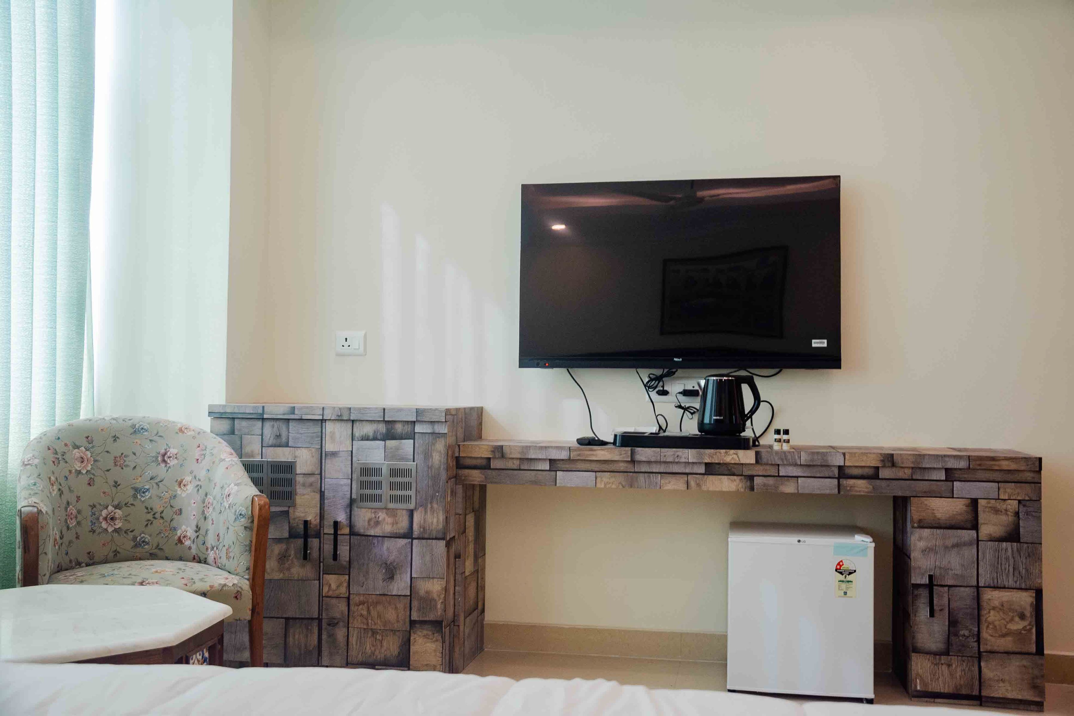 Une chambre dotée d’un téléviseur à écran plat fixé au mur, d’un petit réfrigérateur, d’une armoire en bois et d’un fauteuil à motifs floraux.