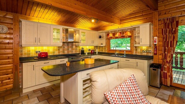 A cozy cabin-style kitchen with wooden beams, a central island, and a window overlooking greenery. - Luxury log cabin with veranda nestled in Kenwick Park, Louth (Louth)