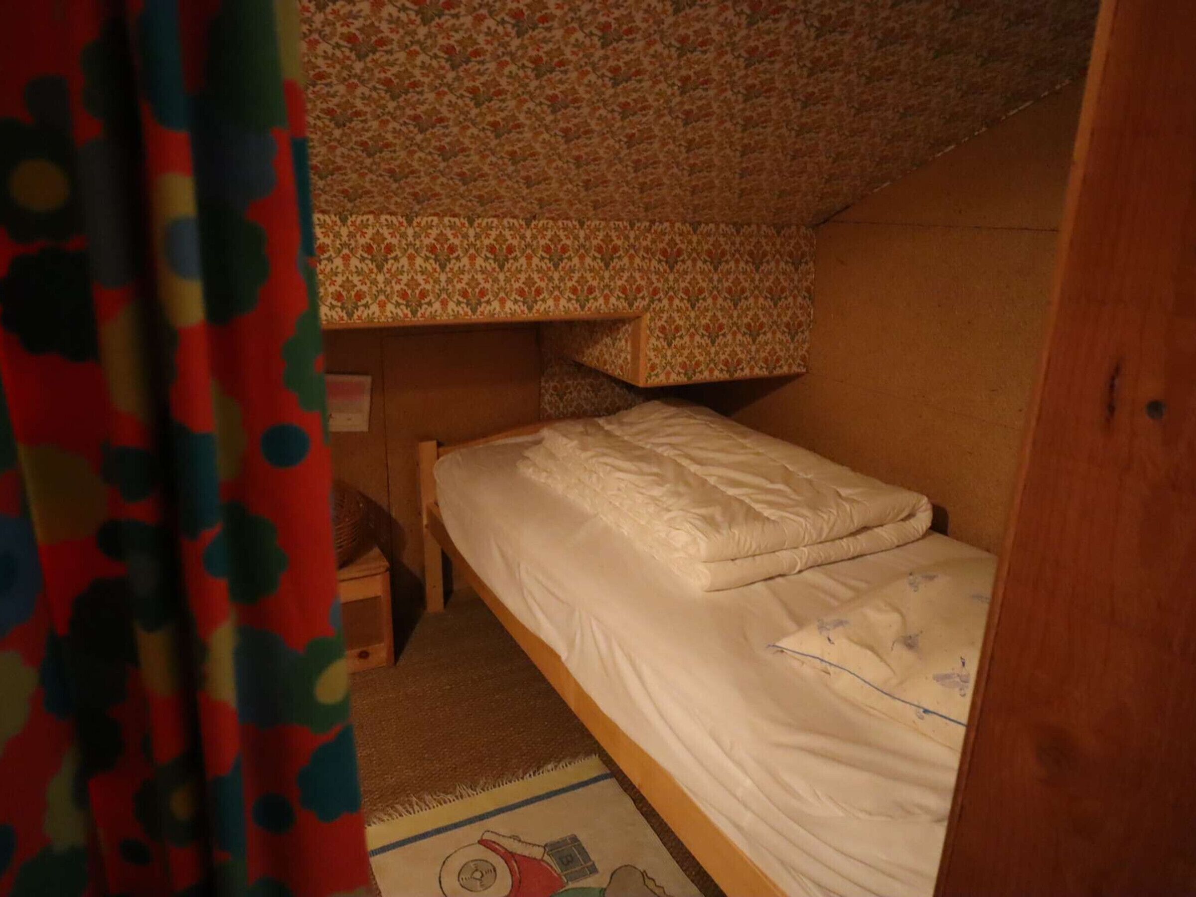 Ein kleines Zimmer mit einem Einzelbett und einem Schlafplatz darüber. Ein kleiner Holzhocker und ein Teppich mit Muster liegen auf dem Boden.
