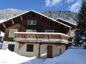 A wooden chalet with a balcony, surrounded by snow-covered trees and a mountain in the background.