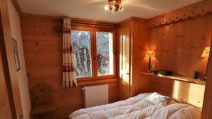 A cozy wooden room with a bed, a window with curtains, a radiator, and a shelf with a lamp. - Chalet for 14 with terrace in the heart of Arêches-Beaufort (Arêches-Beaufort)