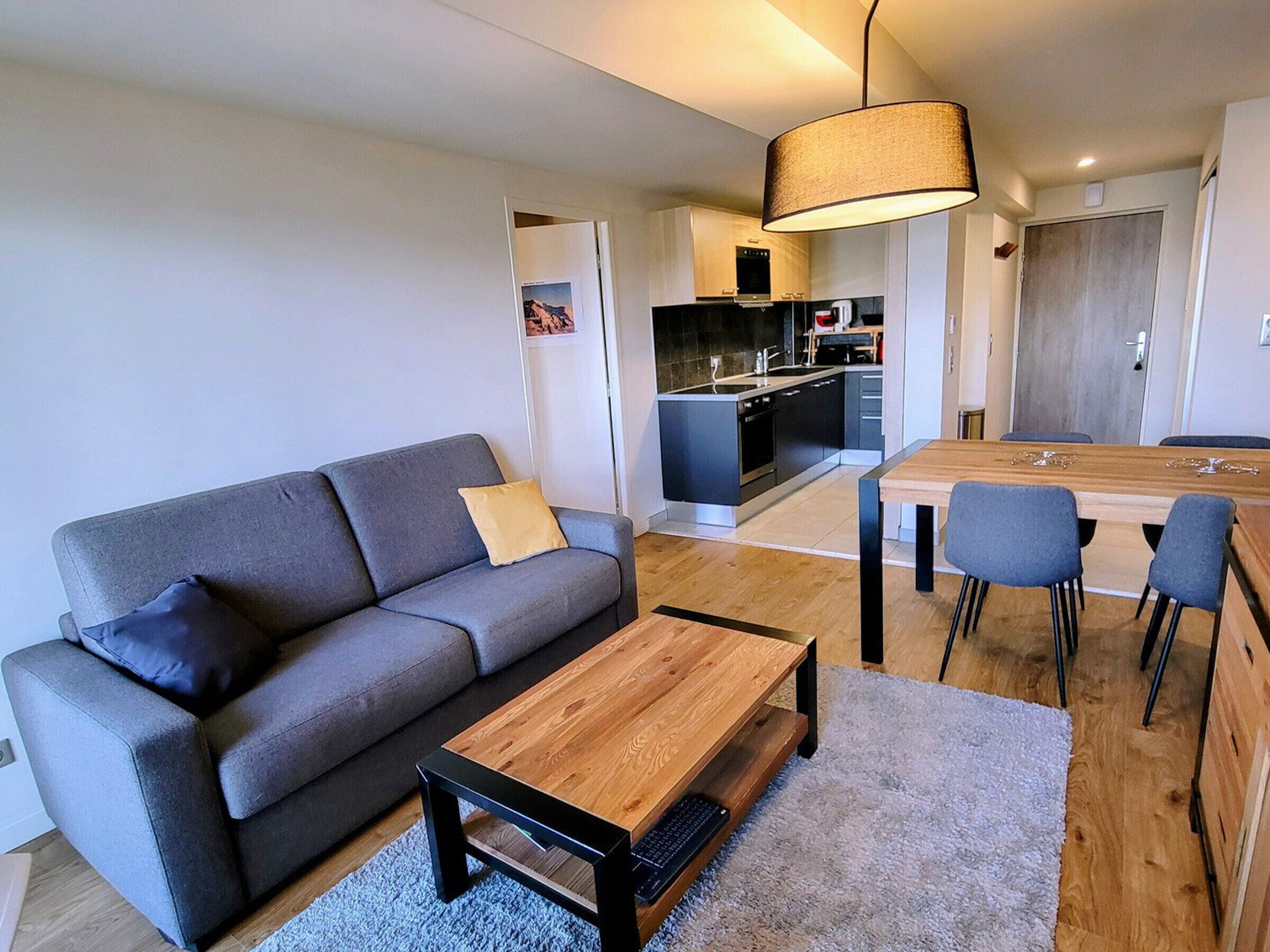 Una sala de estar moderna con un sofá gris, una mesa de centro de madera y una mesa de comedor con sillas. Se ve una cocina con estufa y armarios al fondo.