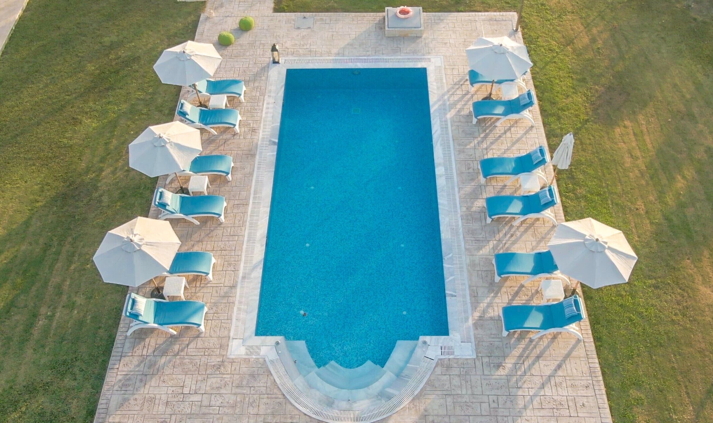 Vue aérienne d’une piscine rectangulaire entourée d’une terrasse carrelée, aménagée avec plusieurs chaises longues et parasols.
