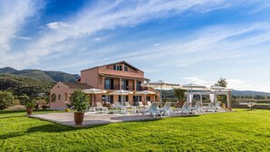 A two-story building with a patio area featuring white lounge chairs and umbrellas, surrounded by a well-maintained lawn and a mountainous backdrop. - Eleni Luxury Villa by White Dream Villas (Corfu)