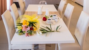 A modern dining area with a white table set for four, surrounded by white chairs. A bouquet of flowers is placed on the table, and there is a television in the background. - Eleni Luxury Villa by White Dream Villas (Corfu)
