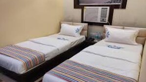 A hotel room with two single beds, each with a striped blanket and a pillow. There is a wall-mounted air conditioner above the beds. - Hotel OPS (Kolkata)