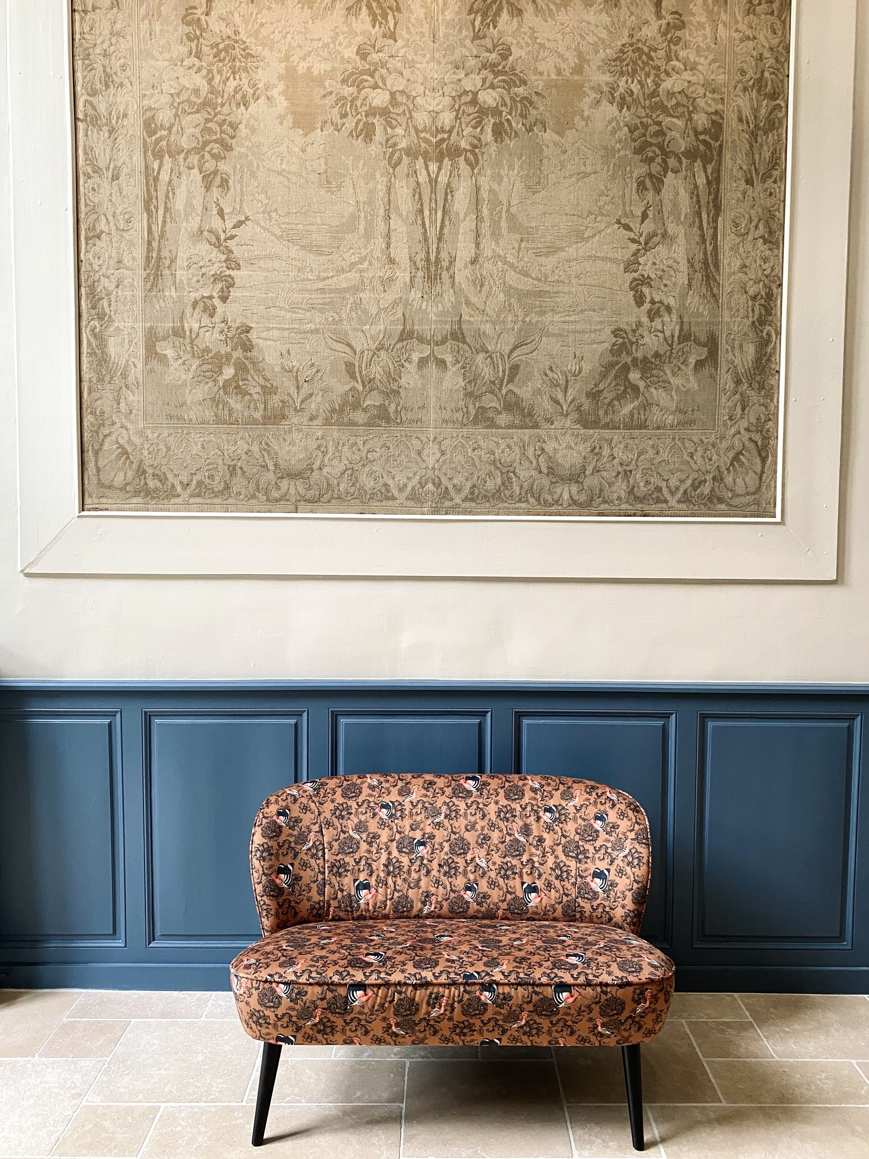 A chair with a floral pattern, a large framed artwork above it, and a blue paneled wall.