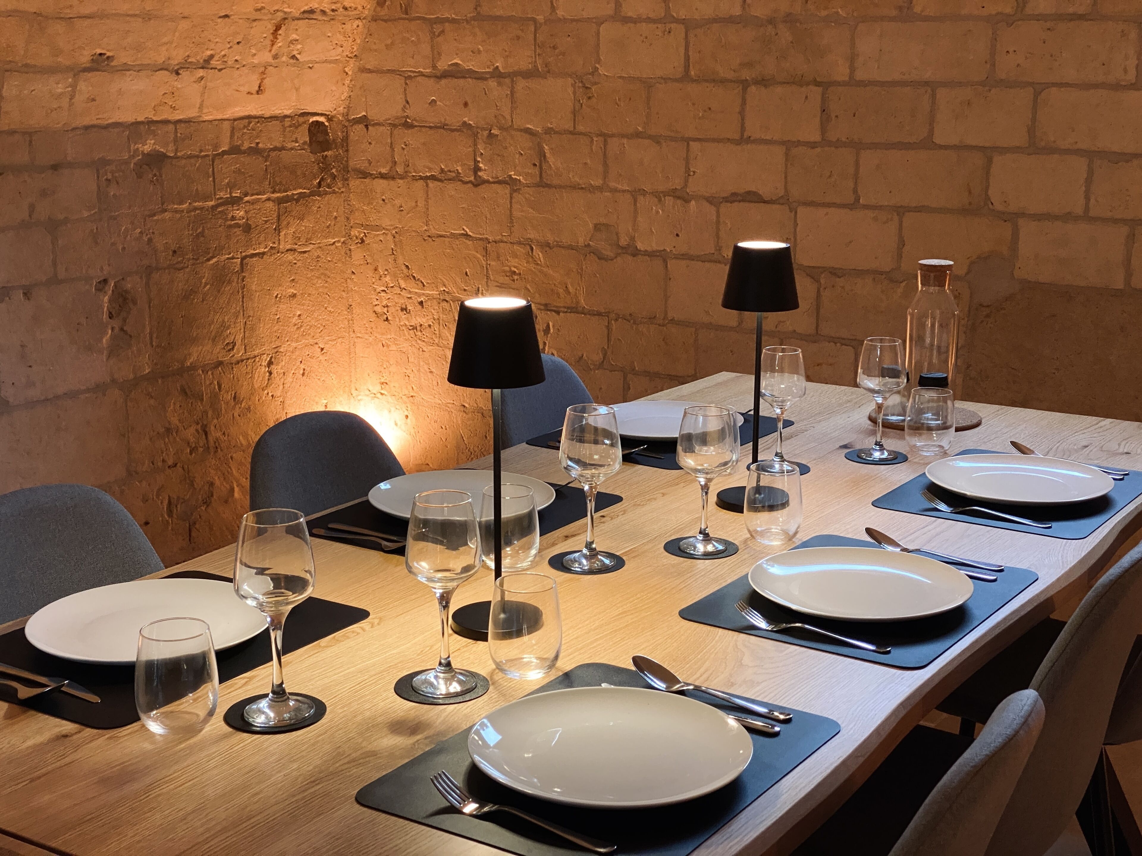 Una mesa de comedor dispuesta con platos, cubiertos y copas de vino, iluminada por lámparas de mesa.