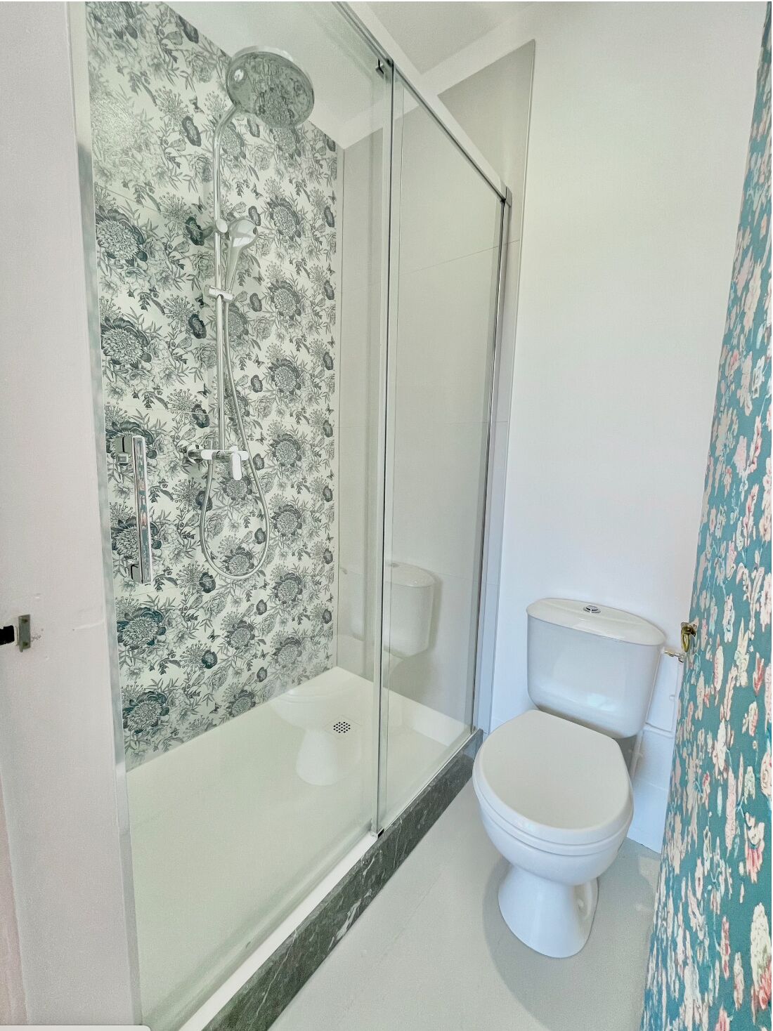 Un baño con ducha envuelta en vidrio, inodoro y papel tapiz estampado con motivos florales.