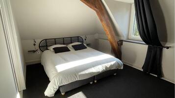 Een slaapkamer met een schuine balklaag, een bed met wit beddengoed, een zwart hoofdeinde en een raam met zwarte gordijnen.