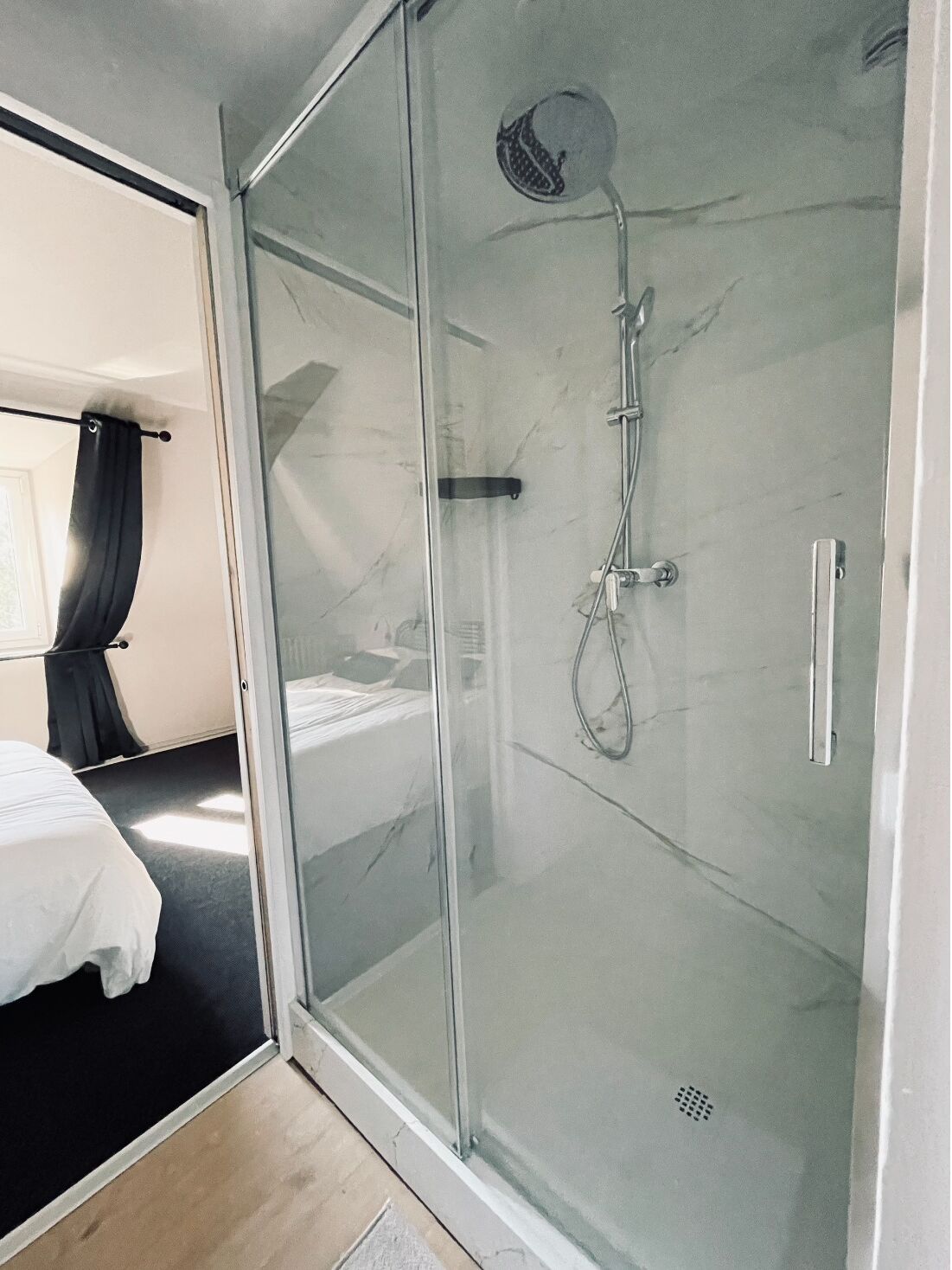 A modern hotel room with a glass-enclosed shower, a bed, and a dark curtain.