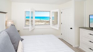 Ein Schlafzimmer mit Bett, Nachttisch, Lampe, Fernseher, Kleiderschrank und Fenster mit Blick auf den Strand.