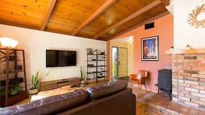 A living room with a wooden ceiling, a brick fireplace, a television, a bookshelf, and a sofa. - 🌲Your Home Base for Exploring the Redwoods! Pet Friendly, Complete with Hot Tub!🐾 (Mckinleyville)
