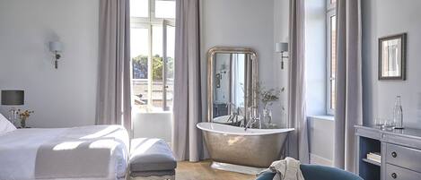 Un dormitorio con cama, una silla, tocador con espejo, bañera y candelabro.
