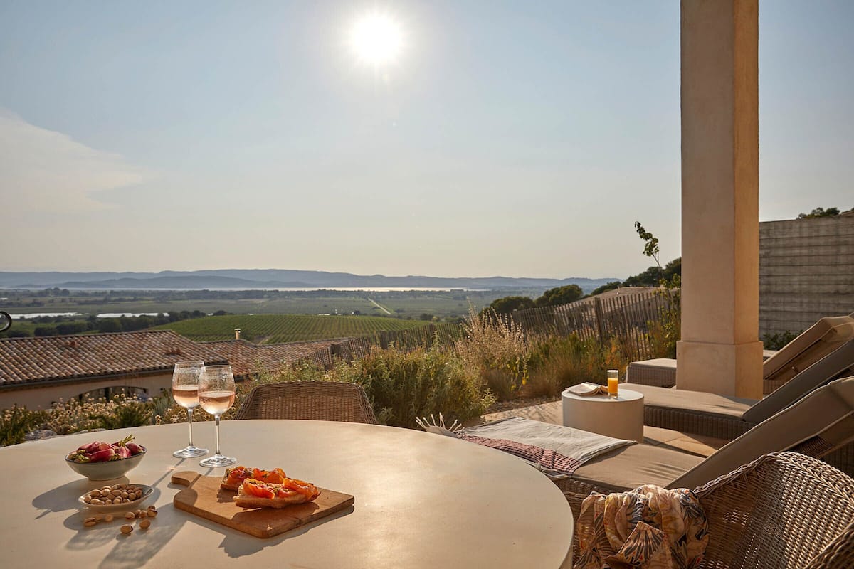 A sunlit terrace with a table set for two, featuring a bowl of nuts, a plate of food, and two glasses of a clear beverage.
