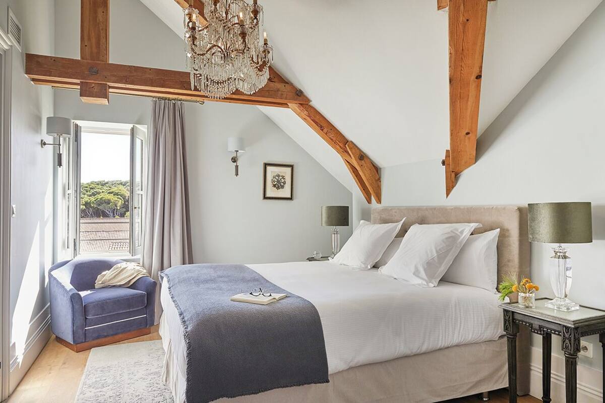 A bedroom with a large bed, a chair, a nightstand, a chandelier, and a view of the outdoors.