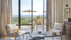 A modern room with a large sliding glass door leading to a balcony with a vineyard view. There are two upholstered chairs, a table with a vase and a glass, and a wooden cabinet. - Château Capitoul (Narbonne)