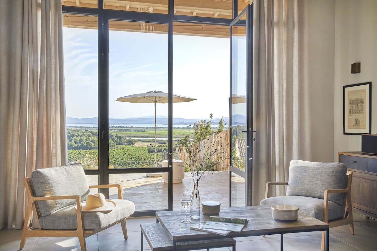 A modern room with a large sliding glass door leading to a balcony with a vineyard view. There are two upholstered chairs, a table with a vase and a glass, and a wooden cabinet.
