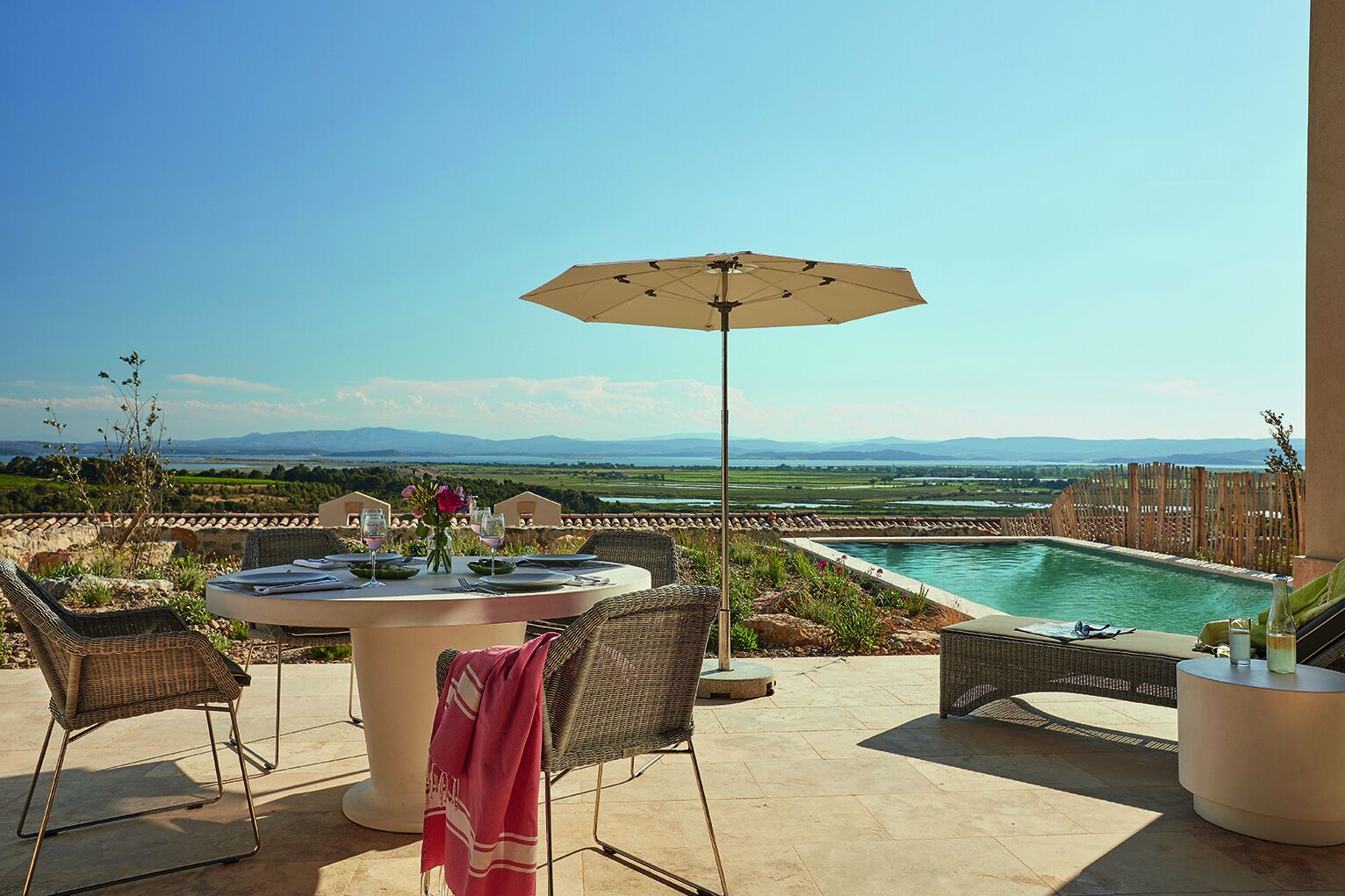 Un patio con una mesa redonda, sillas y una sombrilla con vistas a una piscina y un paisaje pintoresco.