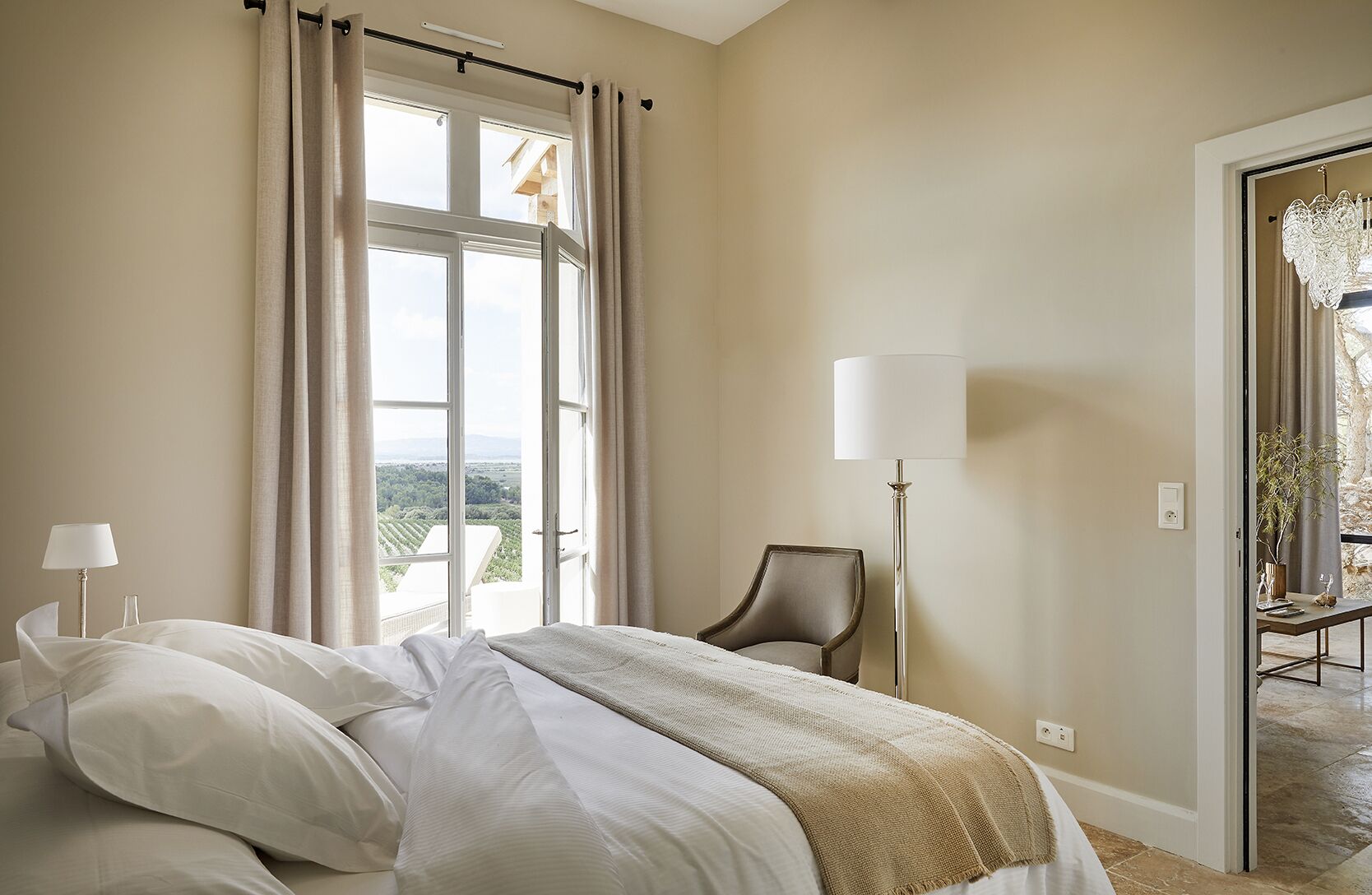 A hotel room with a large bed, a chair, a floor lamp, and a view of the outdoors.