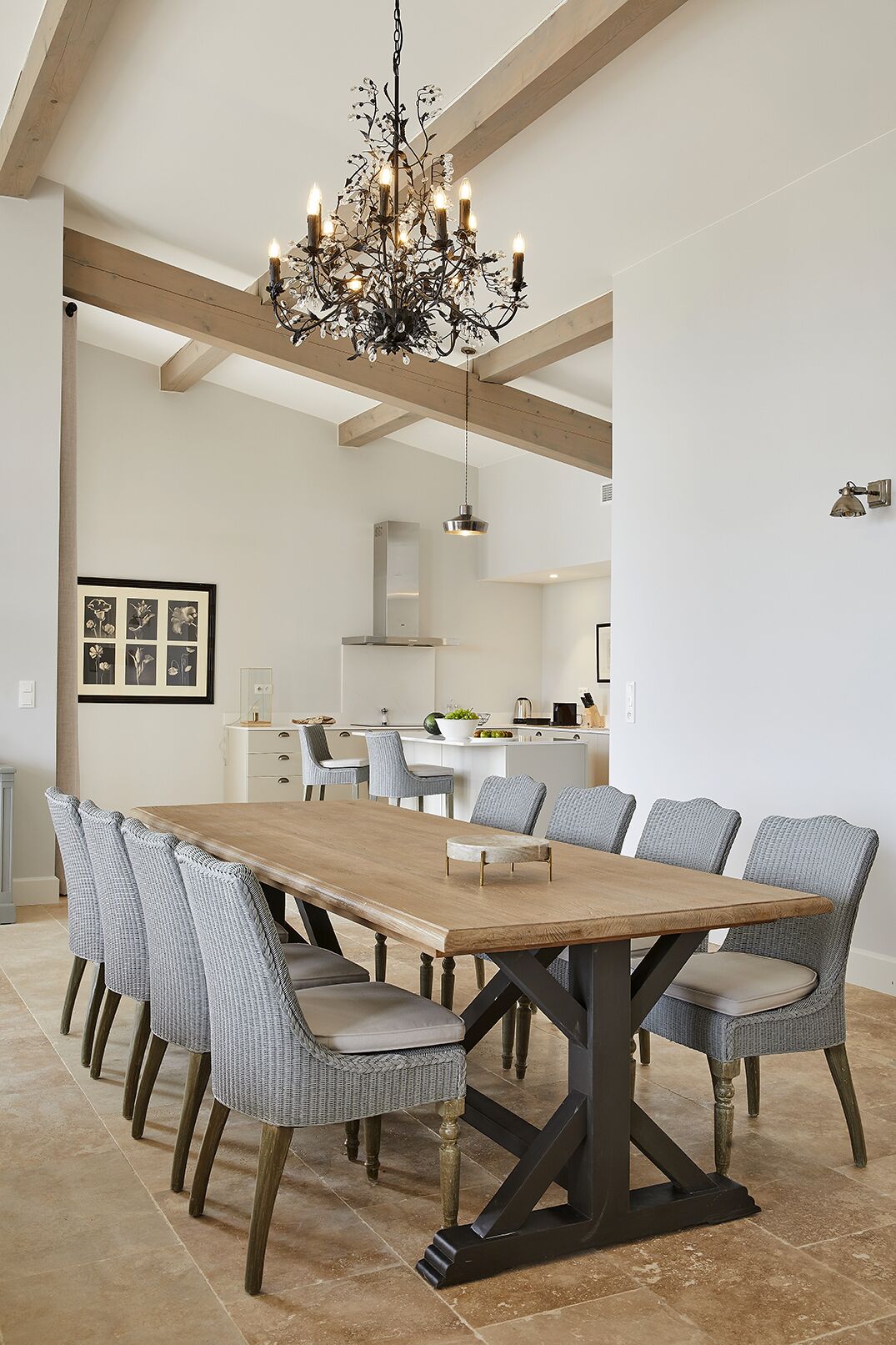 Un espace repas avec une longue table en bois, six chaises rembourrées, un lustre et une cuisine en arrière-plan.
