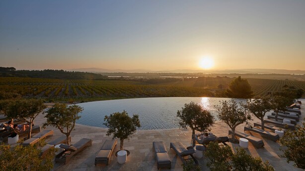 A serene outdoor seating area with lounge chairs and tables overlooking a vineyard and a body of water during sunset. - Château Capitoul (Narbonne)