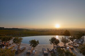 Un espacio tranquilo al aire libre con sillones y mesas con vista a un viñedo y un cuerpo de agua al atardecer.
