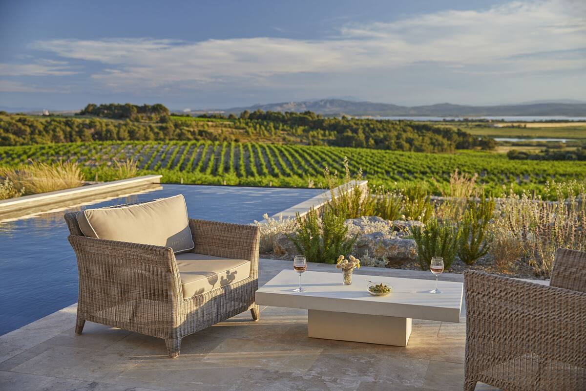 A wicker outdoor seating area with a pool, a white table, and a view of a vineyard.