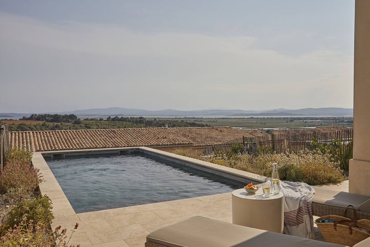 A rectangular swimming pool with a tiled deck, surrounded by outdoor furniture and a view of distant hills.
