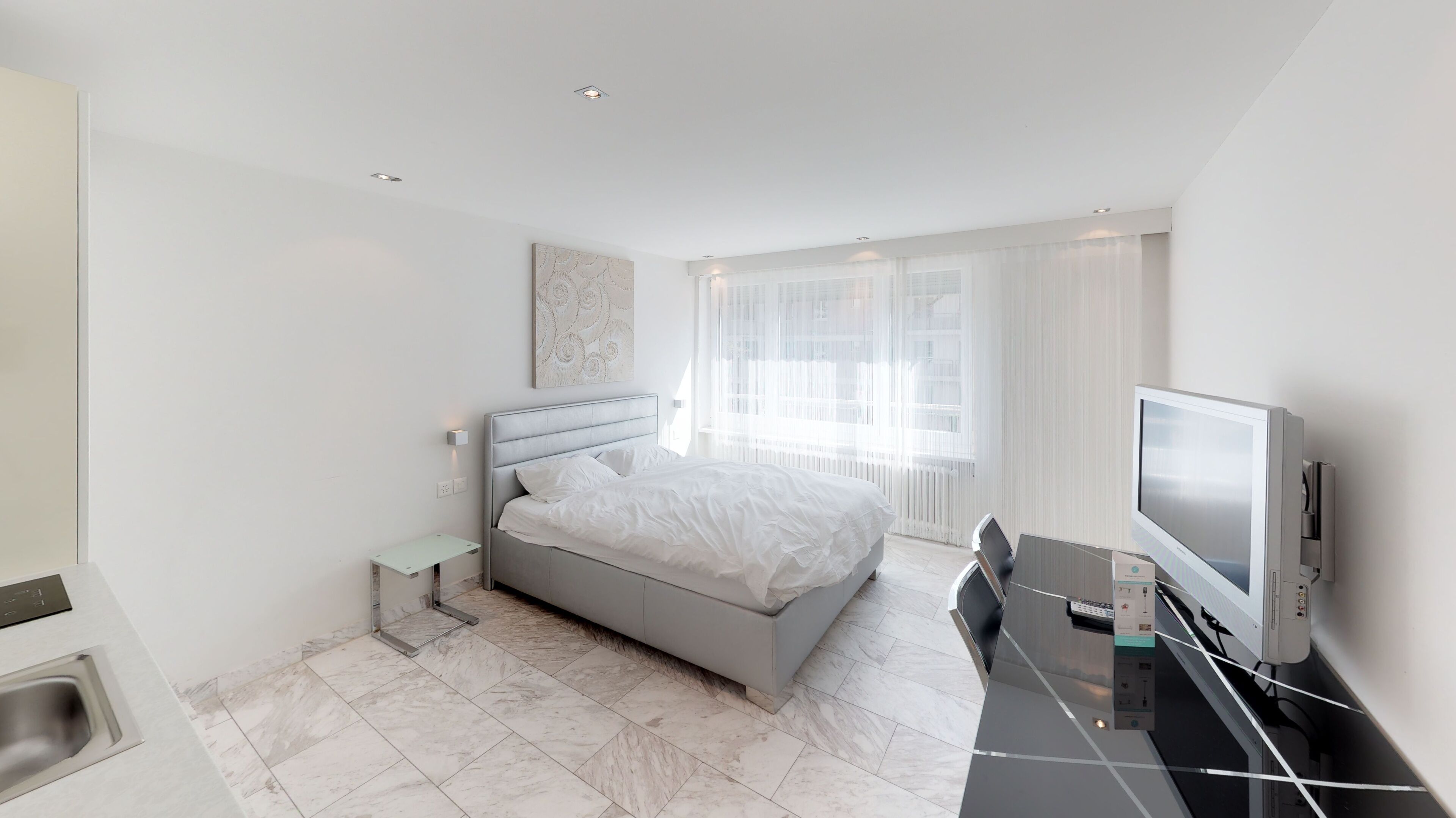 Une chambre d’hôtel moderne équipée d’un lit, d’un bureau avec un téléviseur à écran plat, d’une petite table de chevet et d’une fenêtre avec des rideaux légers.