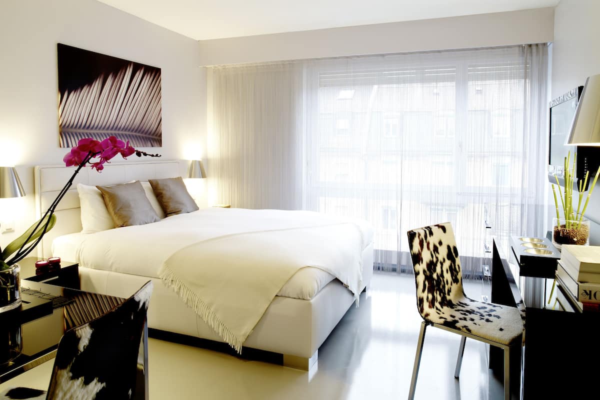 A modern hotel room with a large bed, a desk, a chair, and a window with sheer curtains.