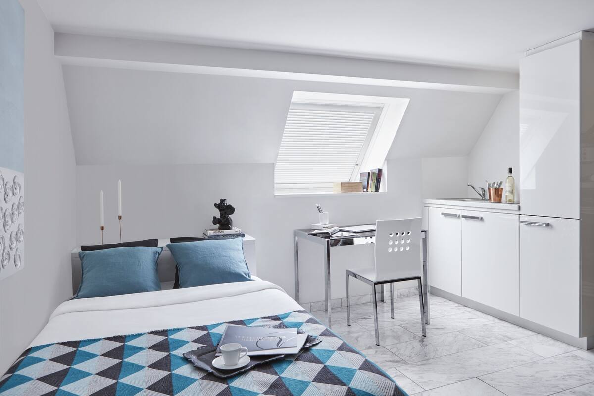 A modern, compact bedroom with a skylight, a neatly made bed, a small desk, and a chair.