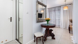 Un pasillo con una puerta blanca, una puerta de vidrio, una mesa redonda de madera con un jarrón de flores amarillas, un pequeño banco acolchado, un espejo con un marco decorativo y una lámpara colgante.
