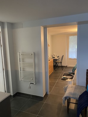 A room with a radiator, a table with a blanket, a chair, and a view into a kitchen area with a window.